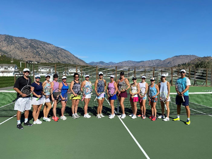 A group of tennis players in colorful clothing pose on the tennis courts. It is bright and sunny out with blue skies.