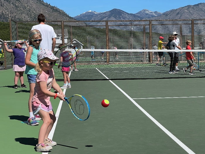 A group of children in colorful outfits hit tennis balls on the tennis court. Youth tennis camps and lessons. Tennis court, tennis players, kids activities.