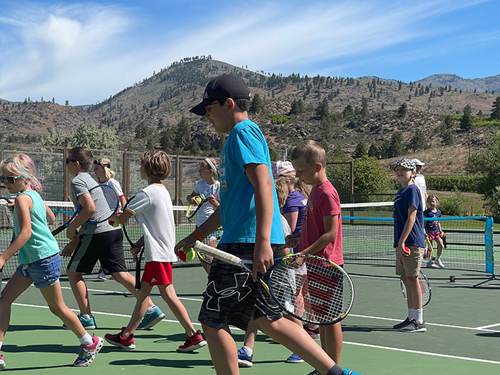 A group of kids walk to the left across the court during a tennis drill. Youth tennis camps, youth tennis lessons.
