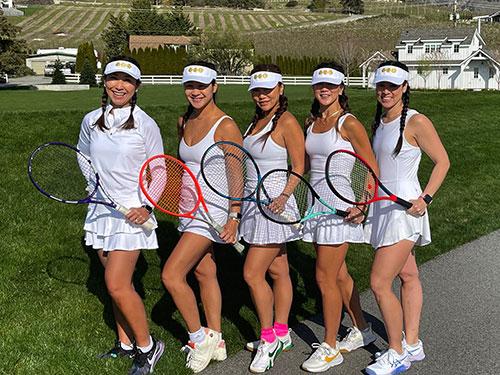 A group of tennis players pose in matching white uniforms holding rackets at their sides. It is bright and sunny with blue skies. Adult tennis camps.
